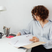 Mujer sentada en su escritorio revisando documentos con concentración, rodeada de materiales de trabajo, representando organización y preparación profesional.