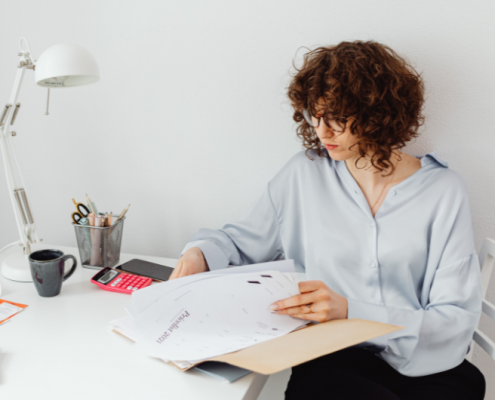 Mujer sentada en su escritorio revisando documentos con concentración, rodeada de materiales de trabajo, representando organización y preparación profesional.
